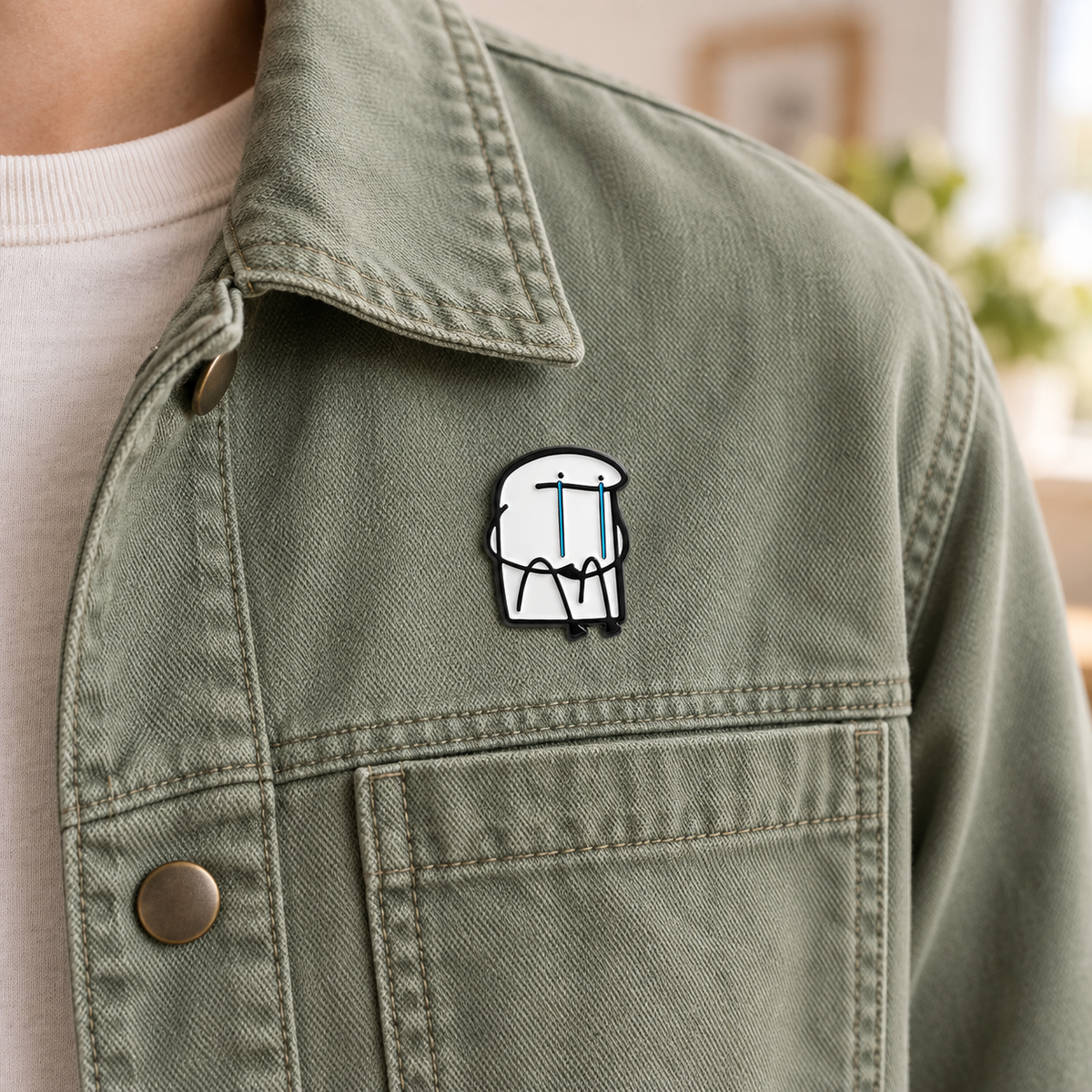 AI-styled wearing view of Sitting Crying Character Pin on muted sage green cotton work jacket being worn by an adult. Pin fixed beside upper chest seam