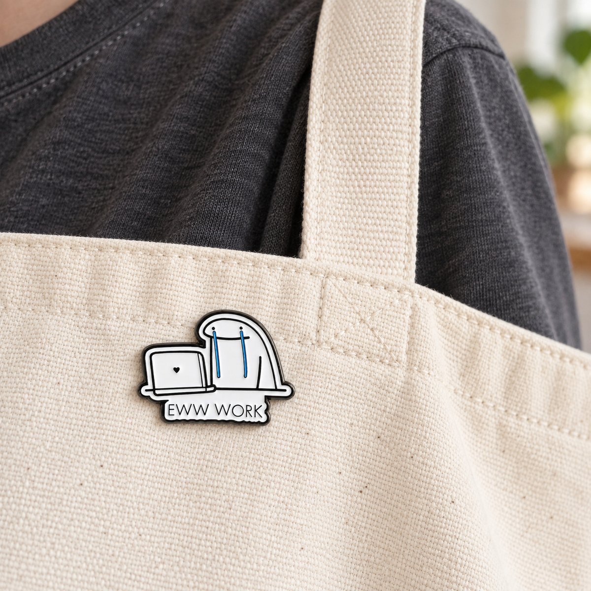 AI-styled wearing view of Eww Work Crying Character Laptop Pin on natural cream canvas tote carried on an adult's shoulder over charcoal cotton shirt. Pin below bag top hem beside 3cm-wide strap. Preserve exact product text EWW WORK legibly