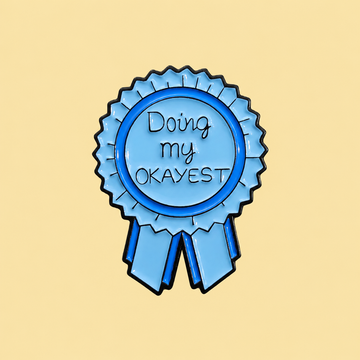 Blue Doing My Okayest award ribbon enamel pin on a pale butter yellow background