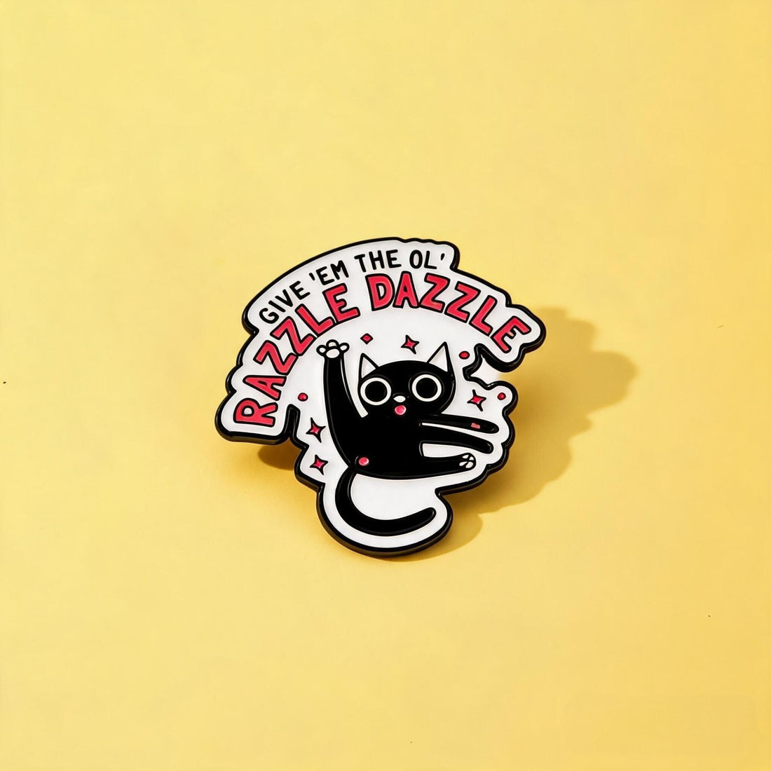Black-and-white cat enamel pin with Give 'Em the Ol' Razzle Dazzle lettering and pink sparkles on yellow
